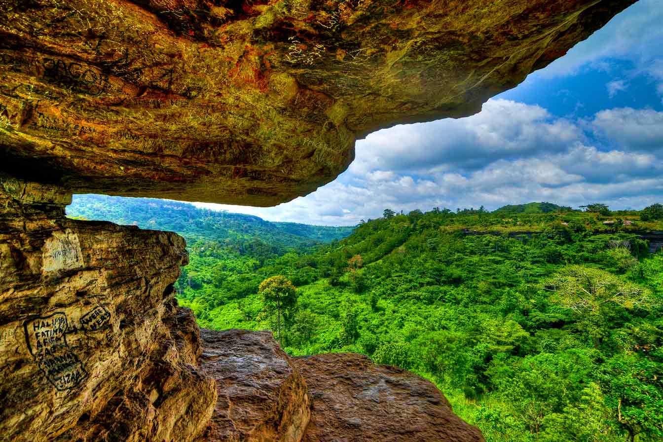 View from moss-covered rock cave overlooking lush green forested valley and hills under blue sky with clouds in Ghana